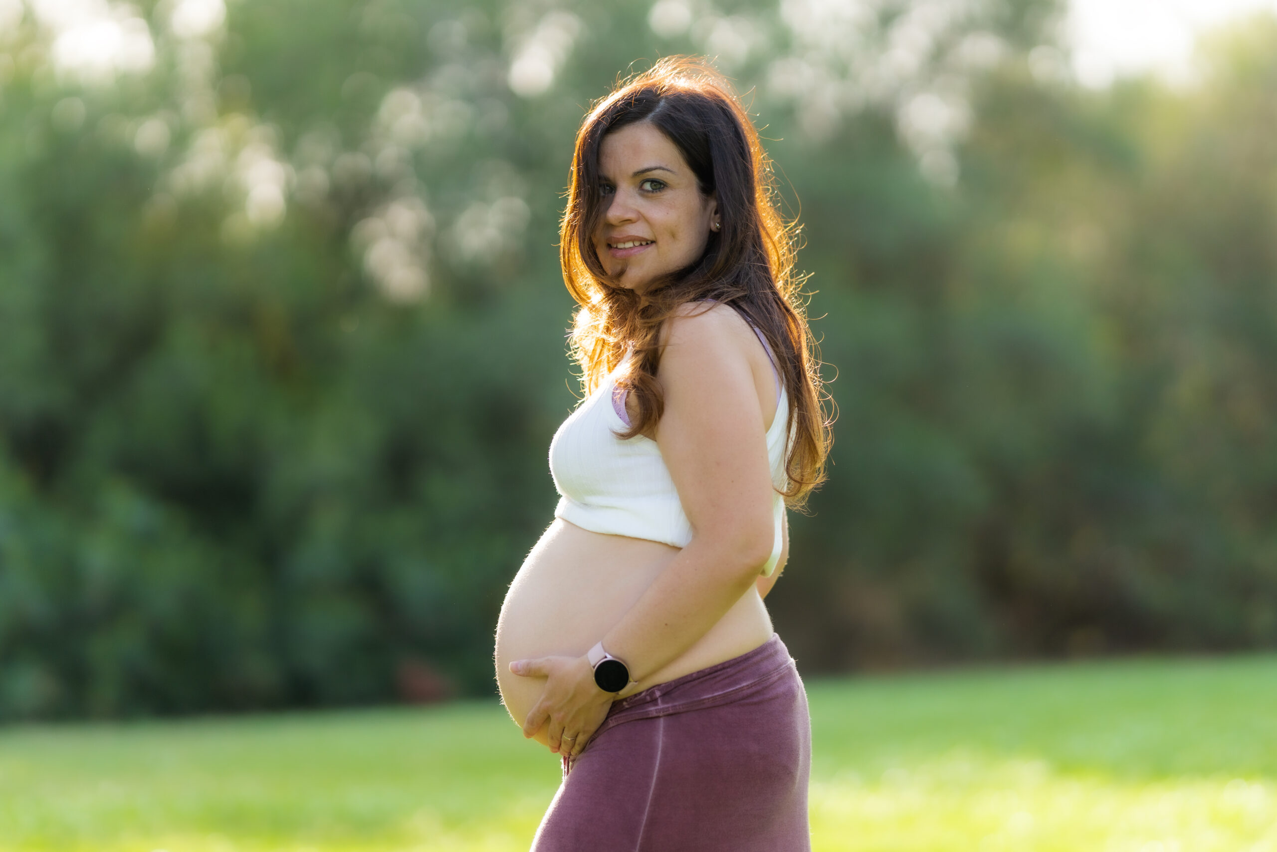 Profile of an adult pregnant latina woman touching her abdomen while facing the camera wearing comfortable sportswear with her abdomen in the air in a park with the sunlight shining on her