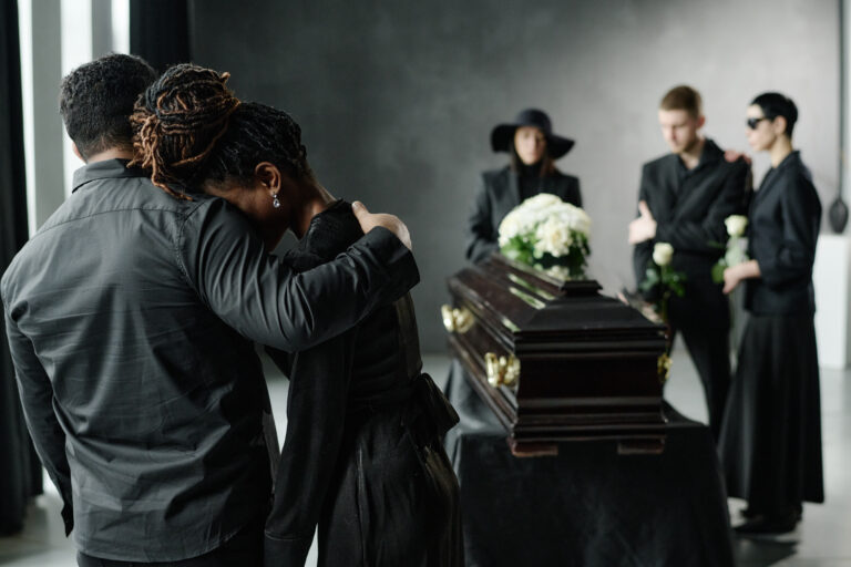 Black woman leaning on Black man for support while grieving near coffin, group of young adults and middle aged woman standing in background holding flowers